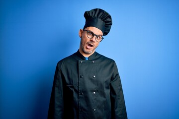 Young handsome chef man wearing cooker uniform and hat over isolated blue background In shock face, looking skeptical and sarcastic, surprised with open mouth