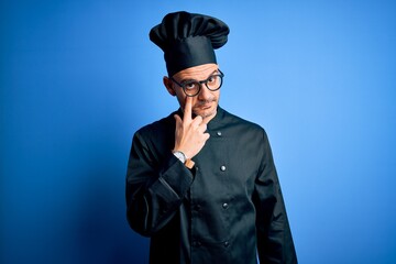 Young handsome chef man wearing cooker uniform and hat over isolated blue background Pointing to the eye watching you gesture, suspicious expression