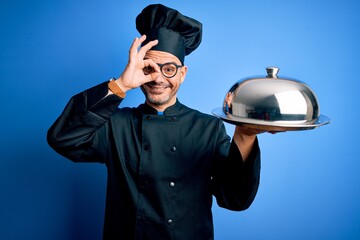 Young handsome chef man wearing uniform and hat holding waiter tray with dome with happy face smiling doing ok sign with hand on eye looking through fingers