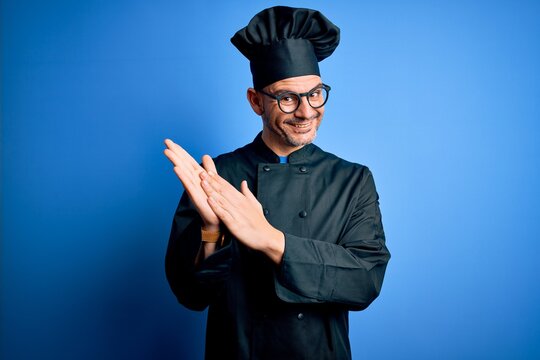 Young handsome chef man wearing cooker uniform and hat over isolated blue background clapping and applauding happy and joyful, smiling proud hands together