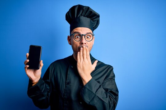 Young handsome chef man wearing uniform and hat holding smartphone over blue background cover mouth with hand shocked with shame for mistake, expression of fear, scared in silence, secret concept