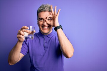 Young modern man drinking a glass of fresh water over purple isolated background with happy face smiling doing ok sign with hand on eye looking through fingers