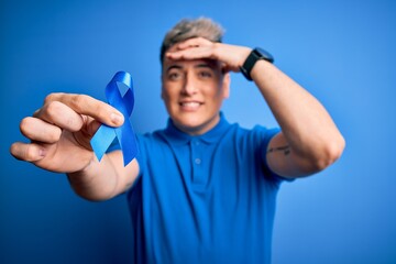 Naklejka premium Close up of young man holding colon cancer awareness blue ribbon over isolated background stressed with hand on head, shocked with shame and surprise face, angry and frustrated. Fear and upset.