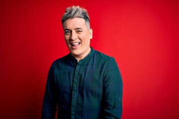 Young handsome modern man wearing elegant green shirt over red isolated background winking looking at the camera with sexy expression, cheerful and happy face.