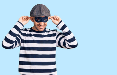 Young handsome man wearing burglar mask smiling pulling ears with fingers, funny gesture. audition problem