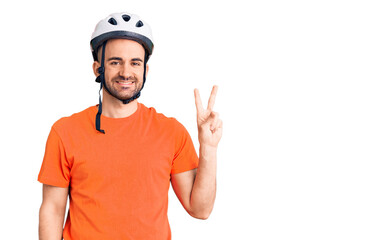 Young handsome man wearing bike helmet smiling with happy face winking at the camera doing victory sign. number two.