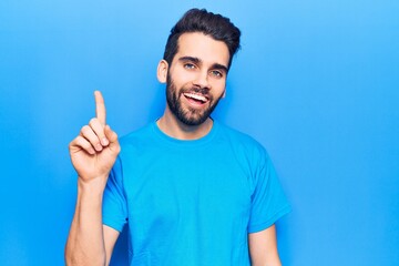 Fototapeta premium Young handsome man with beard wearing casual t-shirt smiling with an idea or question pointing finger up with happy face, number one