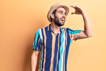 Young handsome man with beard wearing summer hat and shirt very happy and smiling looking far away with hand over head. searching concept.