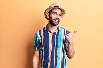 Young handsome man with beard wearing summer hat and shirt smiling with happy face looking and pointing to the side with thumb up.