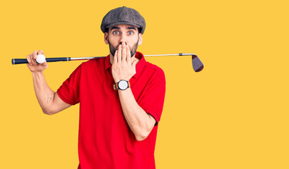 Young handsome man with beard playing golf holding club and ball covering mouth with hand, shocked...