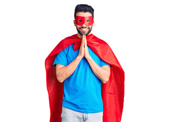 Young handsome man with beard wearing super hero costume praying with hands together asking for forgiveness smiling confident.