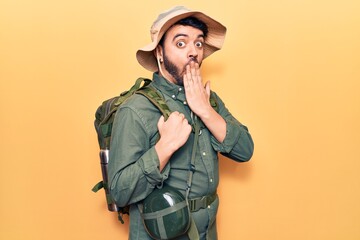 Young hispanic man wearing explorer hat and bakcpack covering mouth with hand, shocked and afraid...