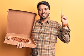 Young hispanic man holding delivery pizza box smiling with an idea or question pointing finger with...