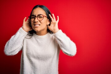 Young beautiful asian woman wearing casual sweater and glasses over red background Trying to hear both hands on ear gesture, curious for gossip. Hearing problem, deaf