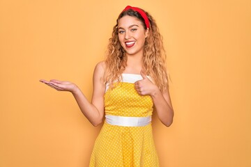 Beautiful blonde pin-up woman with blue eyes wearing diadem standing over yellow background Showing palm hand and doing ok gesture with thumbs up, smiling happy and cheerful