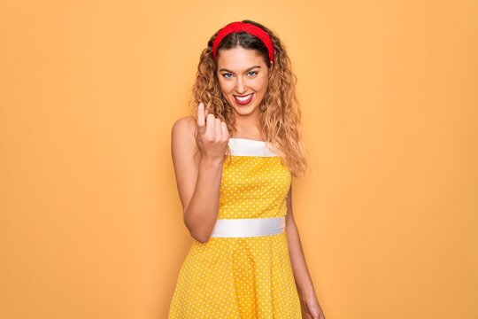 Beautiful blonde pin-up woman with blue eyes wearing diadem standing over yellow background Beckoning come here gesture with hand inviting welcoming happy and smiling