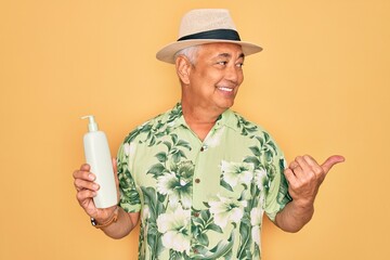 Middle age senior grey-haired man wearing summer hat holding sun protection cream pointing and showing with thumb up to the side with happy face smiling