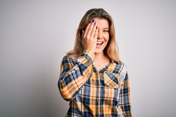 Young beautiful blonde woman wearing casual shirt standing over isolated white background covering one eye with hand, confident smile on face and surprise emotion.