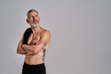 Middle aged muscular man took off black t shirt, showing his body, looking aside while posing in studio over grey background