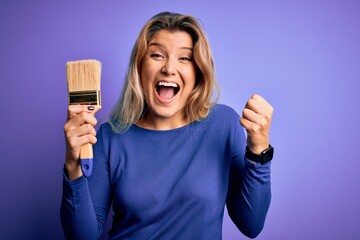 Young beautiful blonde painter woman painting using paint bush over purple background screaming proud and celebrating victory and success very excited, cheering emotion