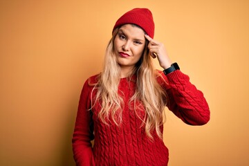 Young beautiful blonde woman wearing casual sweater and wool cap over white background pointing unhappy to pimple on forehead, ugly infection of blackhead. Acne and skin problem