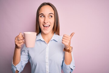 Young beautiful woman drinking mug of coffee standing over isolated pink background pointing and showing with thumb up to the side with happy face smiling