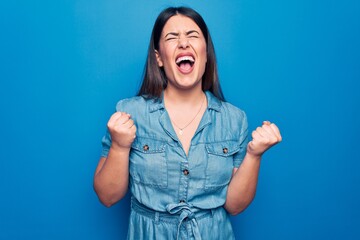 Fototapeta premium Young beautiful brunette woman wearing casual denim dress over isolated blue background celebrating surprised and amazed for success with arms raised and eyes closed