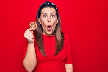 Young beautiful brunette woman holding sweet chocolate cookie over isolated red background scared and amazed with open mouth for surprise, disbelief face