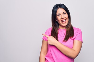 Young beautiful brunette woman wearing casual pink t-shirt standing over white background cheerful with a smile on face pointing with hand and finger up to the side with happy and natural expression