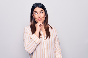 Young beautiful brunette woman wearing casual striped shirt standing over white background thinking concentrated about doubt with finger on chin and looking up wondering