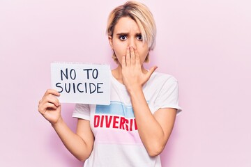 Young beautiful blonde woman wearing t shirt with diversity message holding no to suicide paper covering mouth with hand, shocked and afraid for mistake. surprised expression