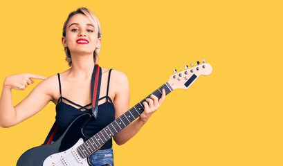 Fototapeta premium Young beautiful blonde woman playing electric guitar pointing finger to one self smiling happy and proud
