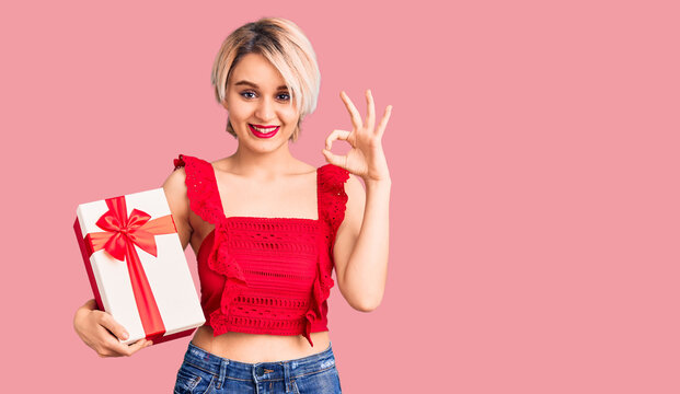 Young beautiful blonde woman holding gift doing ok sign with fingers, smiling friendly gesturing excellent symbol