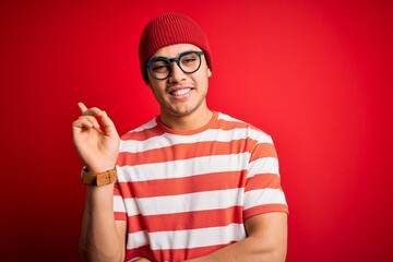 Naklejka premium Young brazilian man wearing casual striped t-shirt and glasses over isolated red background with a big smile on face, pointing with hand and finger to the side looking at the camera.