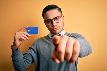 Young brazilian businessman holding credit card money over isolated yellow background pointing with finger to the camera and to you, hand sign, positive and confident gesture from the front