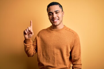 Young brazilian man wearing casual sweater standing over isolated yellow background pointing finger up with successful idea. Exited and happy. Number one.