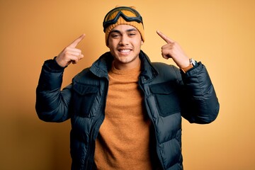 Young brazilian skier man wearing snow sportswear and ski goggles over yellow background smiling pointing to head with both hands finger, great idea or thought, good memory