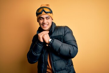 Young brazilian skier man wearing snow sportswear and ski goggles over yellow background laughing...