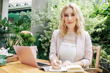 Portrait of young beautiful business lady sitting at desk in her backyard, working on laptop and taking note in planner