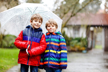 Two little kid boys on way to school walking during sleet, rain and snow with umbrella on cold day....