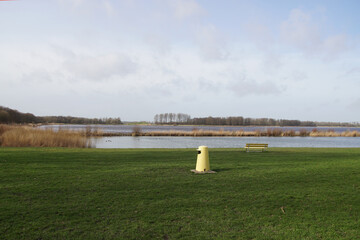 View of the lake from the Geestmerambacht recreation area near the Dutch city of Alkmaar. There is a yellow bench and yellow trash can in the grass on the bank. Netherlands, February 18, 2020.