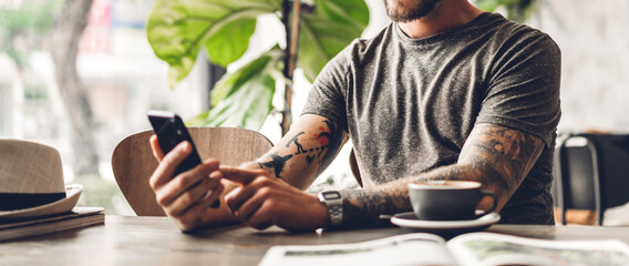 Handsome hipster man relaxing using digital smartphone with coffee and looking at screen typing...