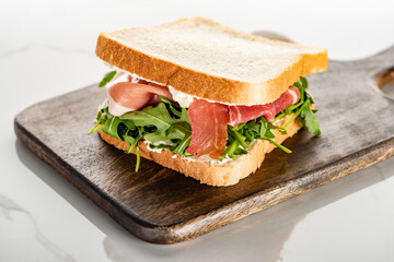 selective focus of fresh green sandwich with arugula and prosciutto on wooden cutting board on white marble surface