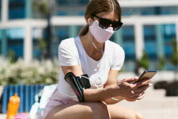 Young fitness woman in sportswear at outdoor sports training. Girl holding a smartphone in her hands and set up music for training. Healthy lifestyle and sports, street training, social distancing