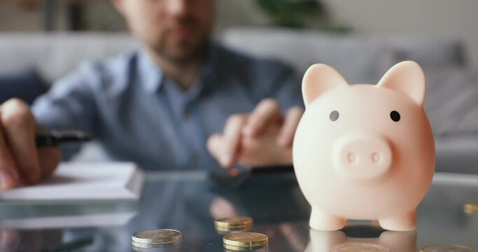 On background man calculates writing in daily planner personal expenses and incomes, close up focus on piggy bank euro coins on table, symbol of budget managing, make savings and take care of tomorrow