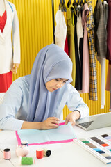 A young beautiful Asian Muslim woman designer who wore a hijab as a startup business owner is sewing the colorful fabric on her working table.