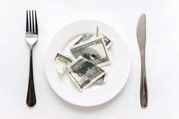 A torn hundred-dollar bill lies on a white plate between a fork and a knife on a yellow tablecloth. The concept of business lunch, banking, life rentier and financial well-being.