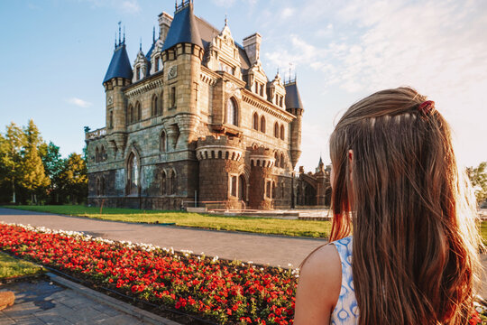 A little girl child in a dress holding a book and stands against the background of a medieval castle, the photo was taken from under a flowerbed