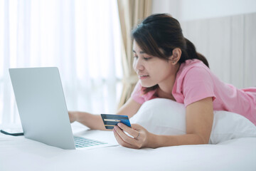 Happy woman holding credit card and using computer laptop for online shopping while making orders on bed in morning at home. Business, technology, ecommerce, digital banking and online payment concept