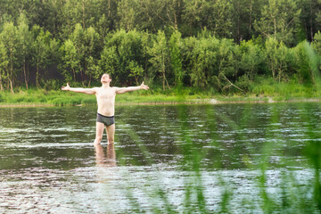 Fototapeta na wymiar A young man stands in the river and with outstretched arms enjoys nature